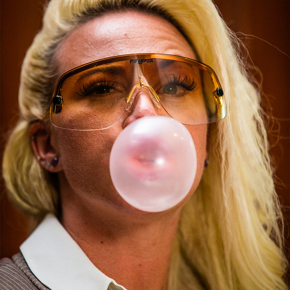 Blonde Frau mit Sonnenbrille und Kaugummi-Blase vor dem Gesicht. Gold Standard Look.