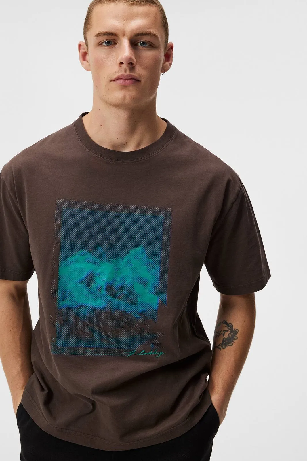 Man wearing a brown T-shirt with a blue-green print. The print is an abstract pattern with the lettering "J.Lindeberg".
