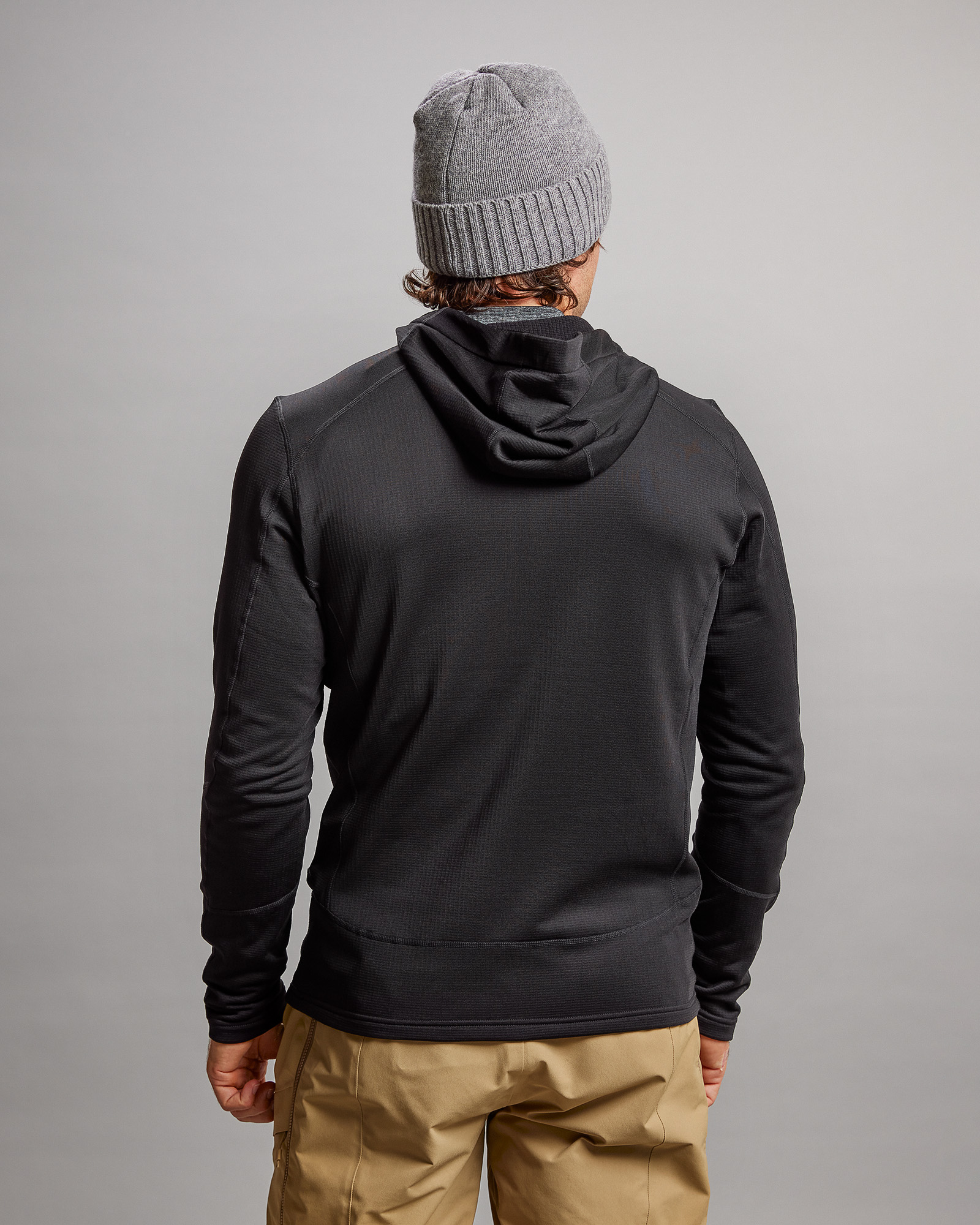 Rückansicht einer schwarzen M-4 Techno Stretch Mid Hood Jacke mit Kapuze, kombiniert mit einer grauen Mütze und beigen Hosen.
