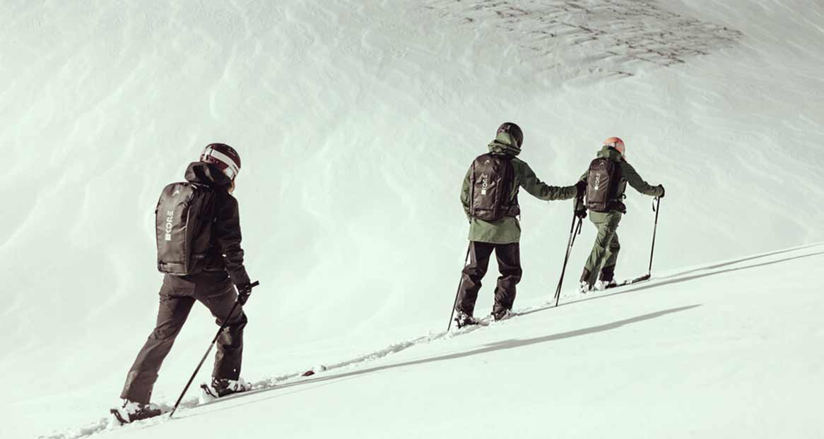 Drei Personen beim Skitourengehen, von hinten gesehen, mit Skiern und Stöcken auf einer verschneiten Piste. Sie tragen Helme und Rucksäcke.