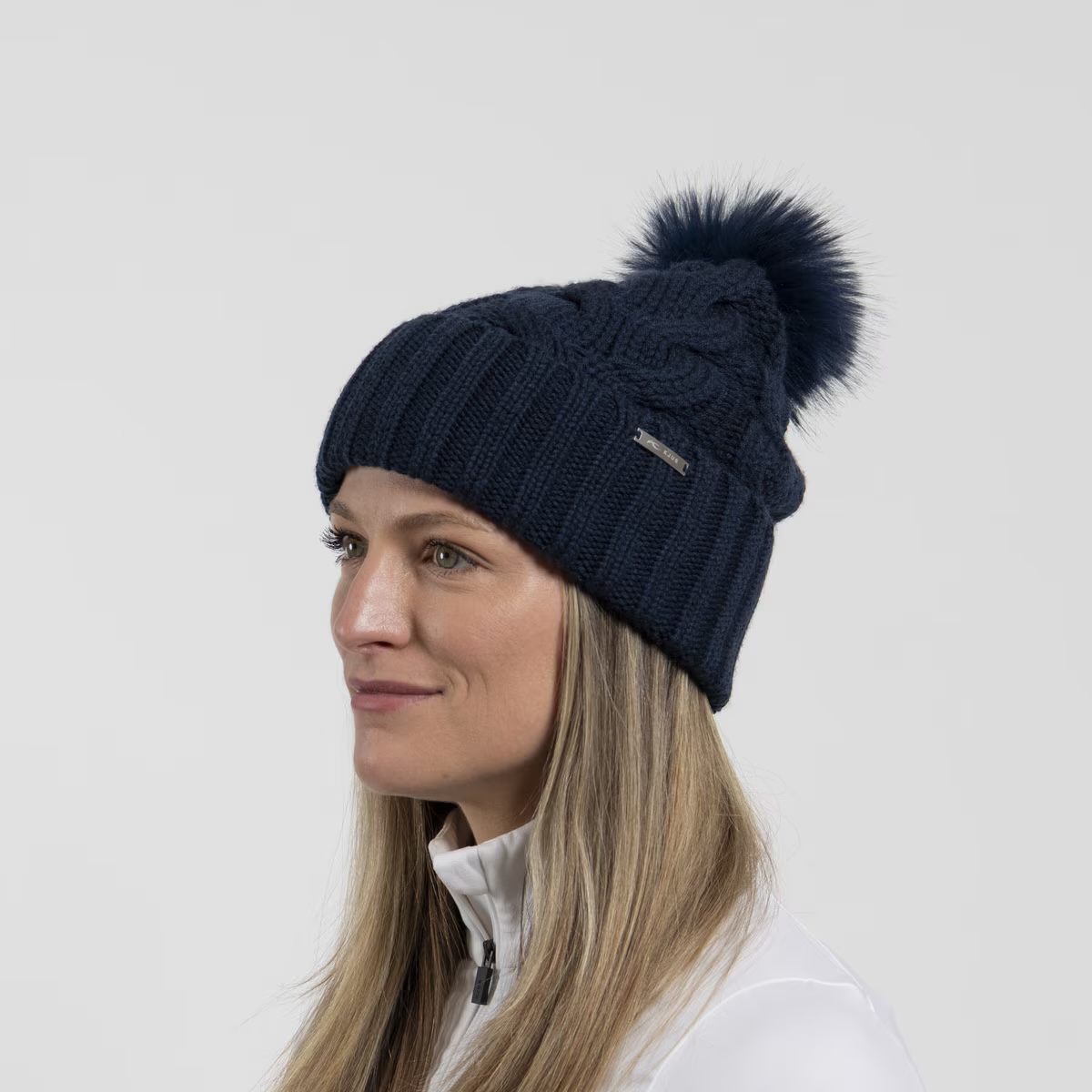 Une femme aux longs cheveux blonds porte un bonnet tricoté bleu foncé avec un pompon et des torsades. Elle regarde vers la gauche.