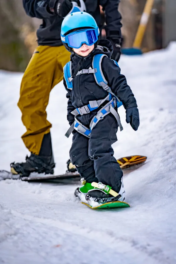 Child with blue helmet and ski goggles, with "The One" snowboard harness, learning to snowboard.