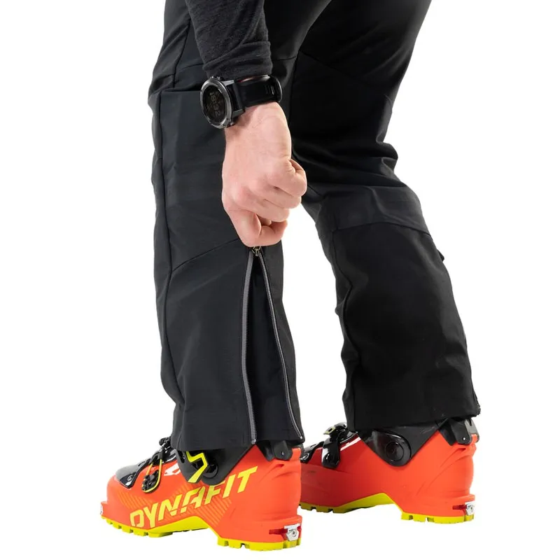 Gros plan : une personne ouvre la fermeture éclair d'un pantalon Radical Infinium Hybrid noir par-dessus une chaussure de ski.