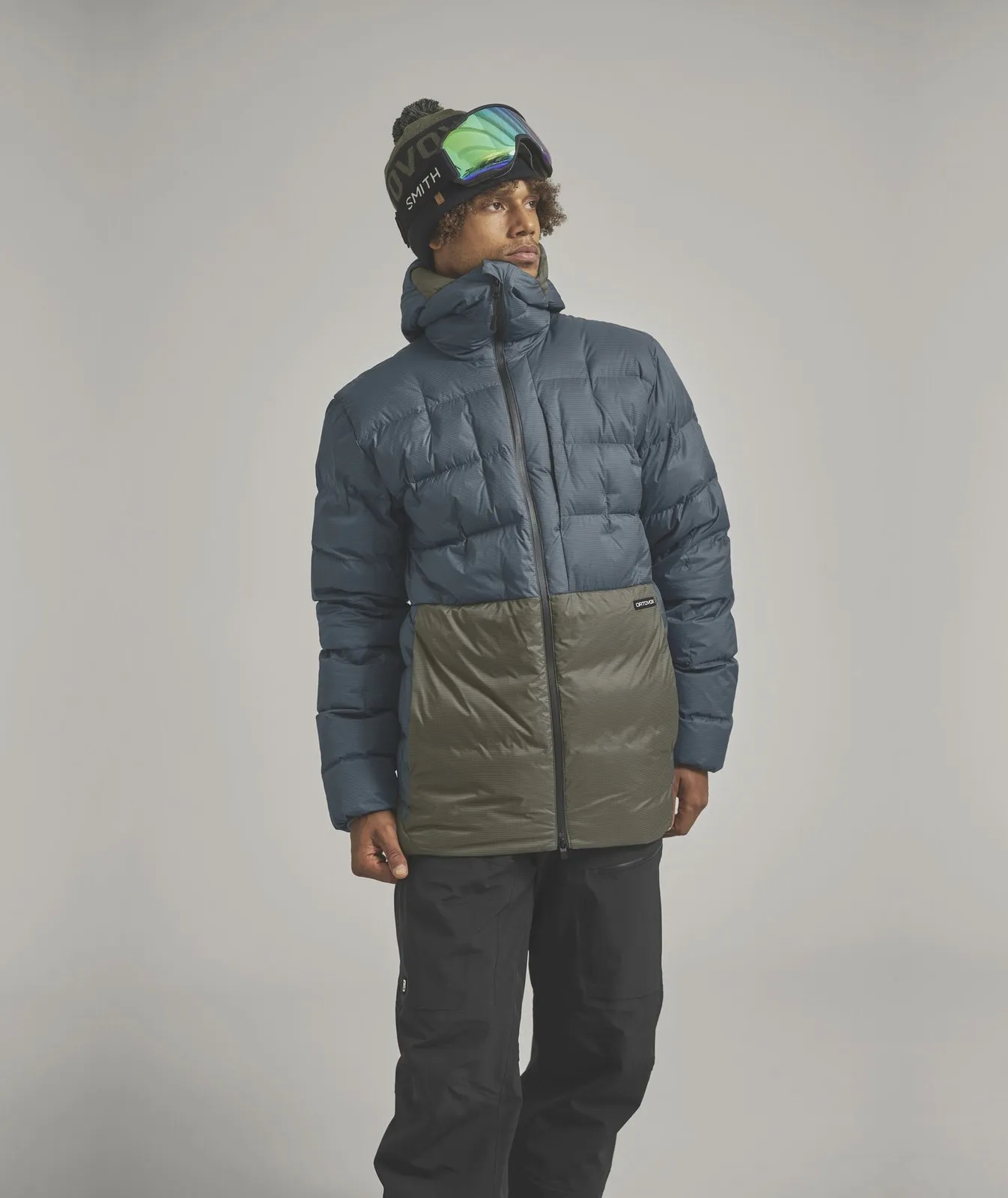 Un uomo con occhiali da sci e berretto indossa un piumino bicolore blu e verde e pantaloni da sci neri.