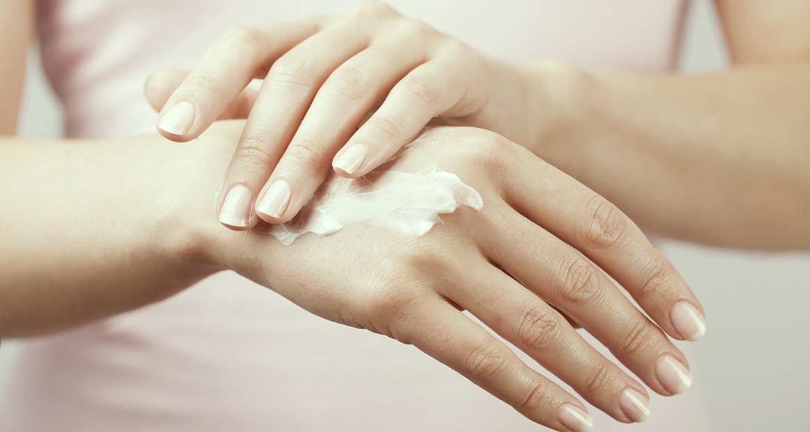 Mains d'une personne qui s'applique de la crème pour les mains sur le dos des mains. Les ongles sont soignés.