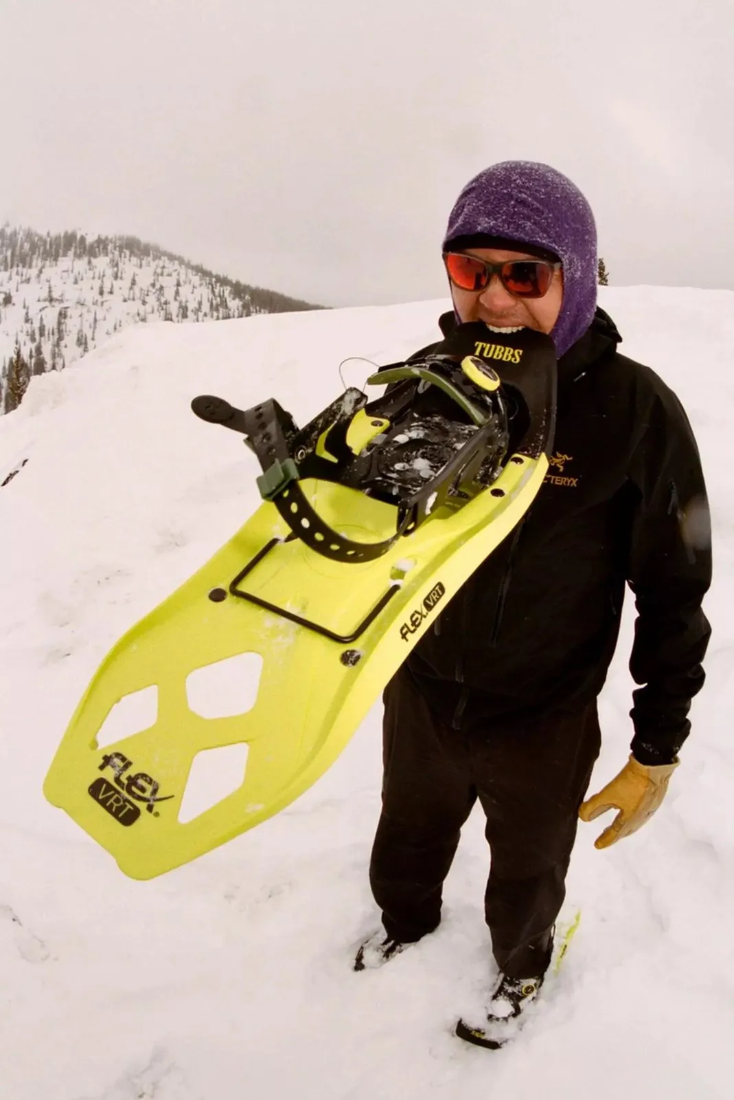 A man holds a yellow Tubbs Flex VRT snowshoe in his mouth, standing in the snow with mountains in the background.