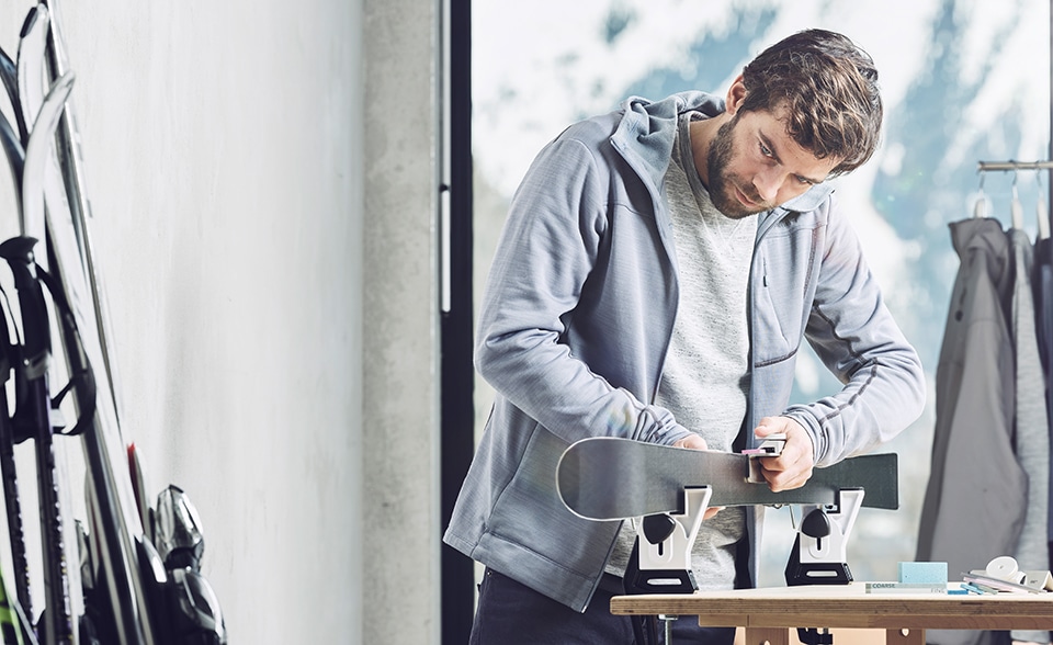 Ein Mann mit Bart schleift eine Skikante in einer Werkstatt, Ski stehen an der Wand.