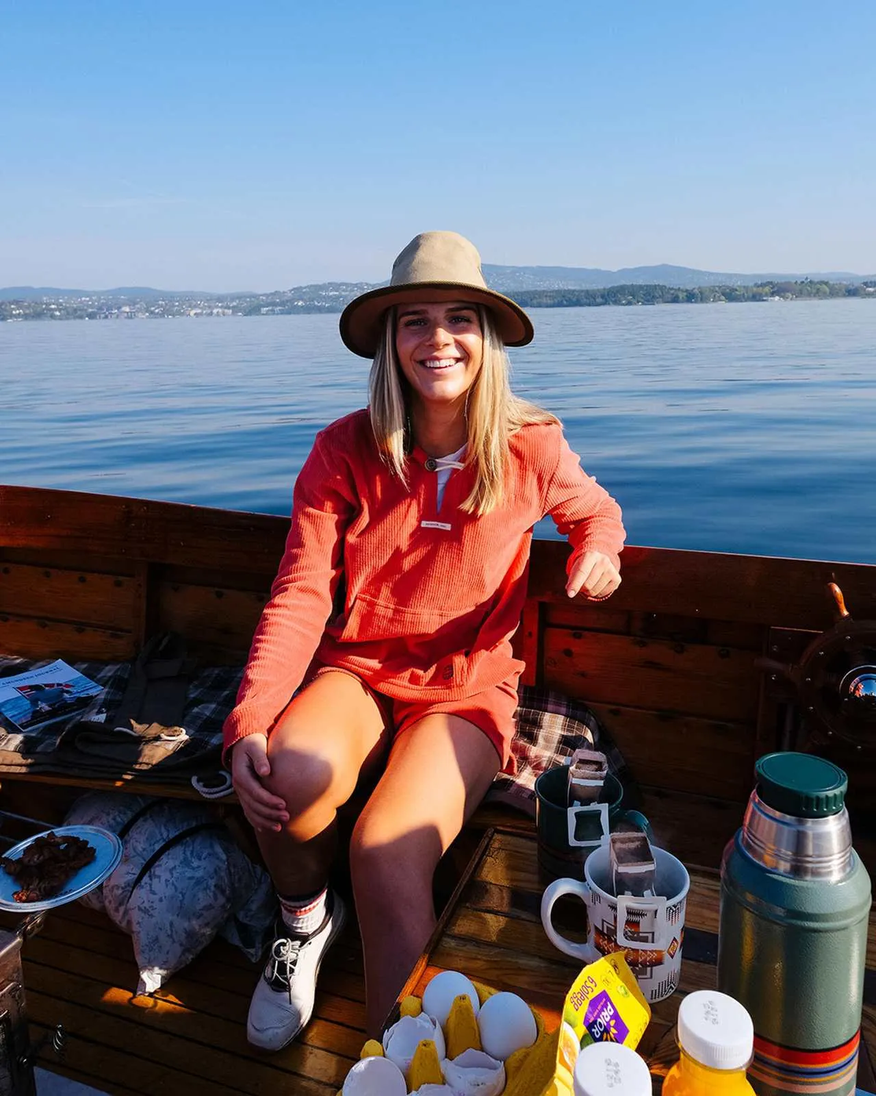 Una donna sorridente con un cappello siede su una barca di legno di fronte all'acqua blu e alle montagne, circondata da cibo e bevande.
