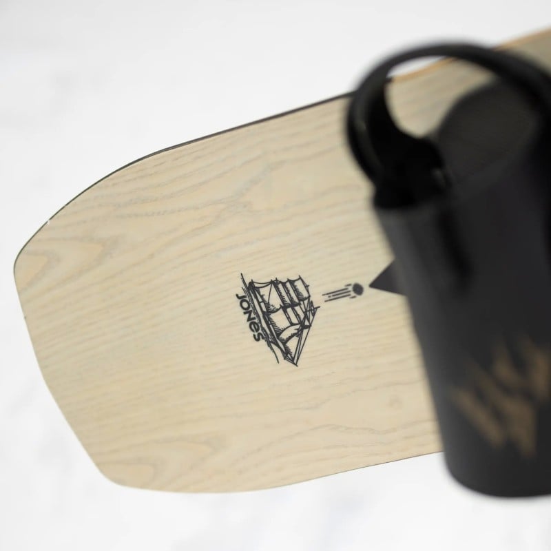 Jones Flagship Pro Snowboard Detail des Jones Flagship Pro Snowboards mit Schiffslogo und Bindung, helles Holzfurnier.