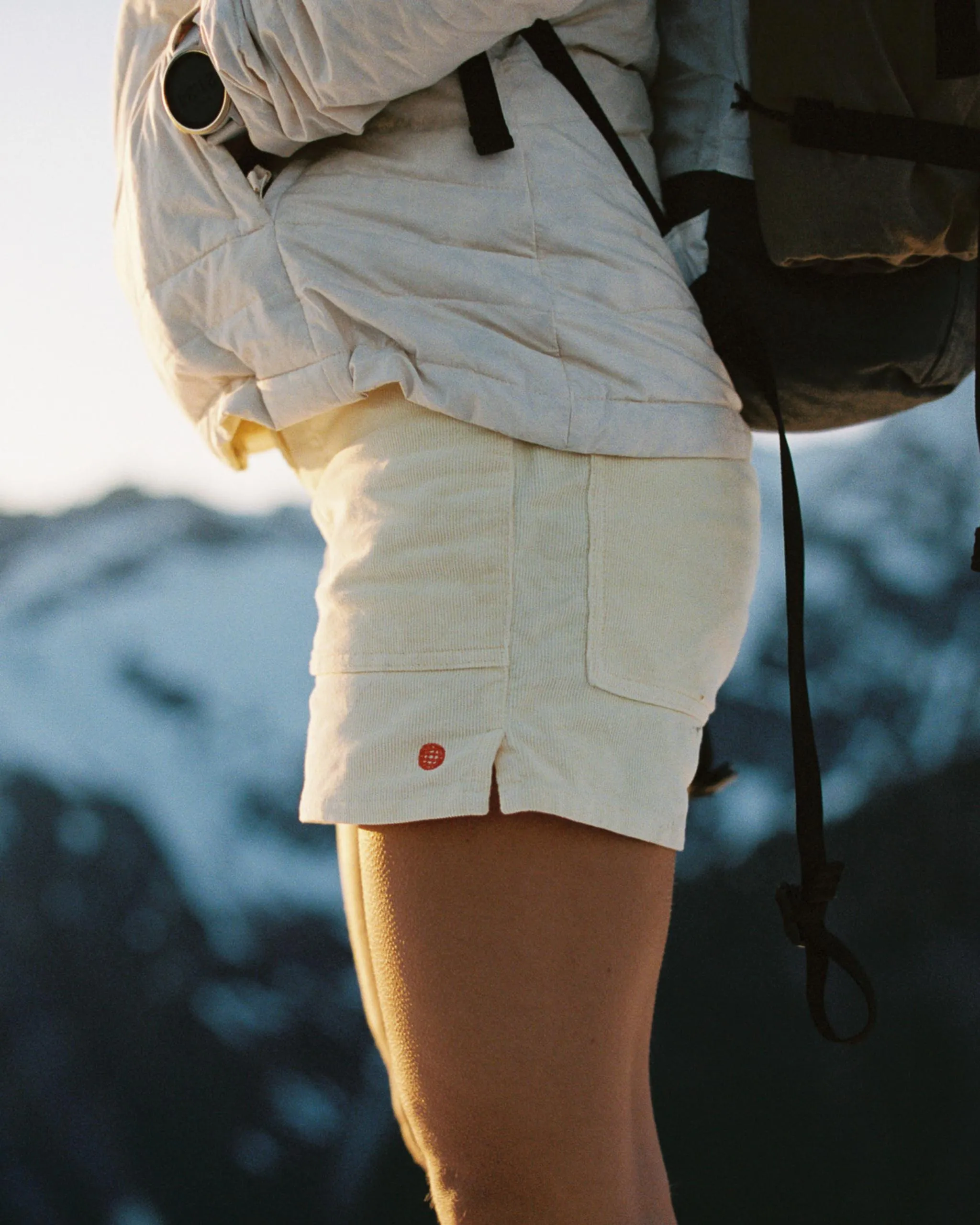 Personne en short et veste avec sac à dos devant un décor de montagne. Petit logo sur le short. La lumière est chaude et dorée.