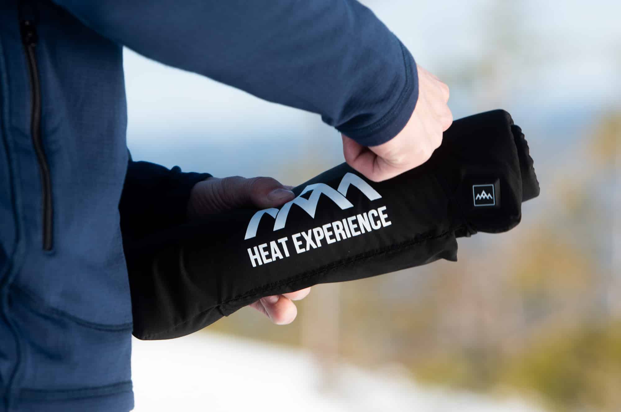 Eine Person hält ein schwarzes Heat Experience Sitzkissen mit weißem Logo in der Hand.