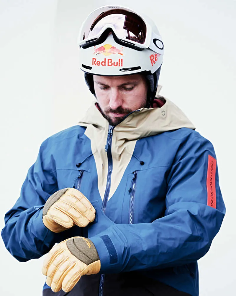Un uomo in tenuta da sci, con casco e occhiali, indossa guanti di pelle. La giacca è blu con un inserto beige.