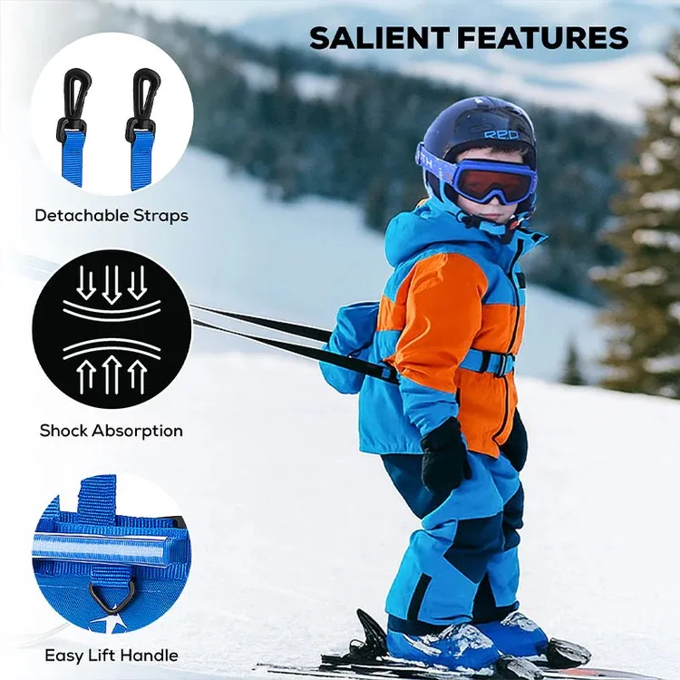 A toddler in orange-and-blue ski clothing, a helmet, and ski goggles stands on skis on a snowy slope. The child is wearing a blue ski harness with black straps on the back. Details: removable straps, padding, lifting handle.