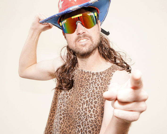 Mann mit Hut, Leopardentop, langen Haaren und polarisierter Sonnenbrille zeigt mit dem Finger.