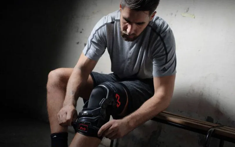 A young man sits on a bench and attaches a black knee brace with red lettering to his right leg.