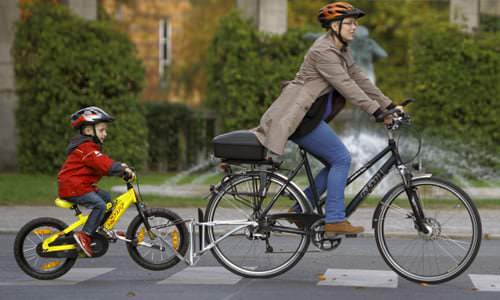 Frau mit Kind auf Fahrrädern, verbunden durch die FollowMe Tandemkupplung. Beide tragen Helme und fahren auf einem Zebrastreifen.