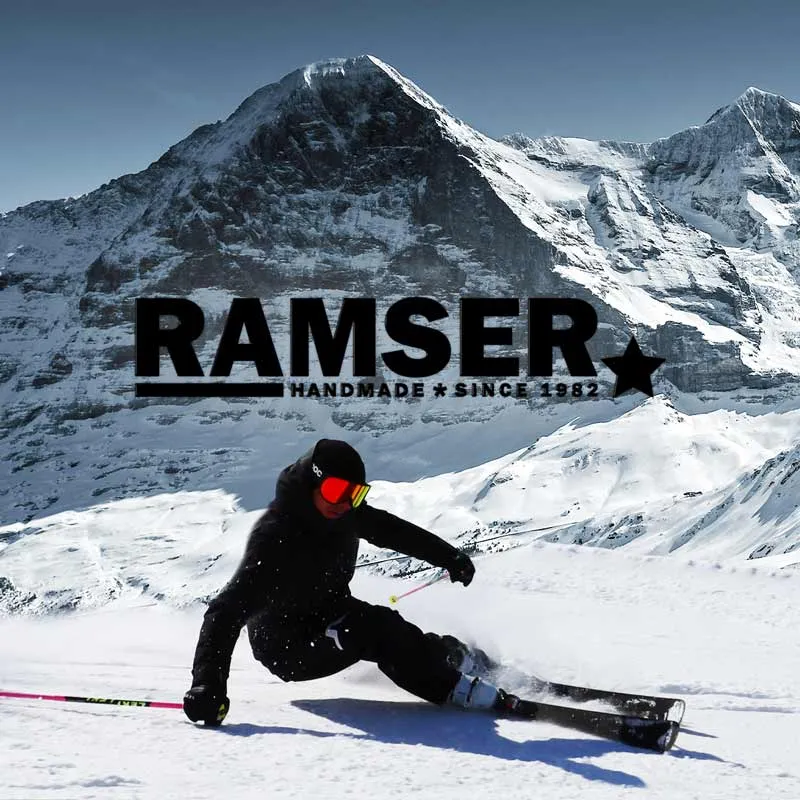 Ein Skifahrer in schwarzer Kleidung fährt vor einer verschneiten Bergkulisse Ski; Text "RAMSER HANDMADE SINCE 1982" im Vordergrund.