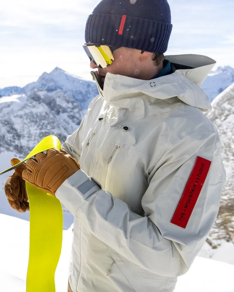 A man in Z-2 HD GORE-TEX Pro 3L Shell Jacket "Castle Wall" with ski goggles and cap holding ski skins in front of snowy mountains.