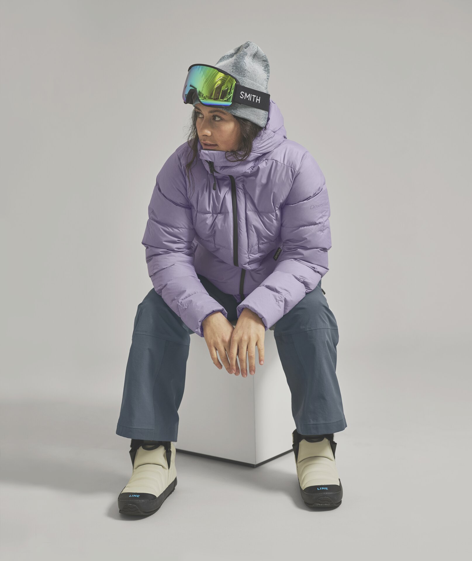 Eine Frau mit Mütze und Skibrille, trägt eine lila Daunenjacke, graue Hose und cremefarbene Stiefel, während sie auf einem weißen Würfel sitzt.