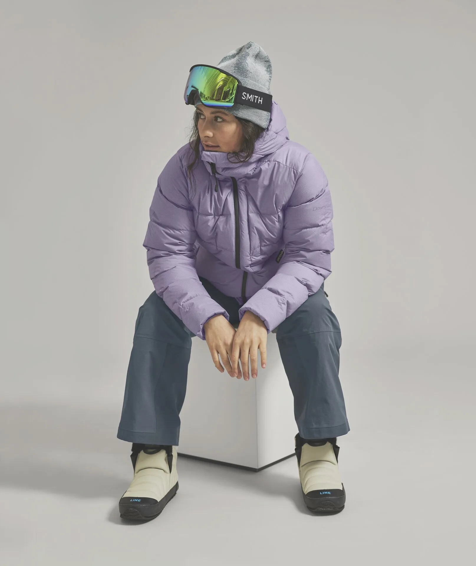 Eine Frau mit Mütze und Skibrille, trägt eine lila Daunenjacke, graue Hose und cremefarbene Stiefel, während sie auf einem weißen Würfel sitzt.