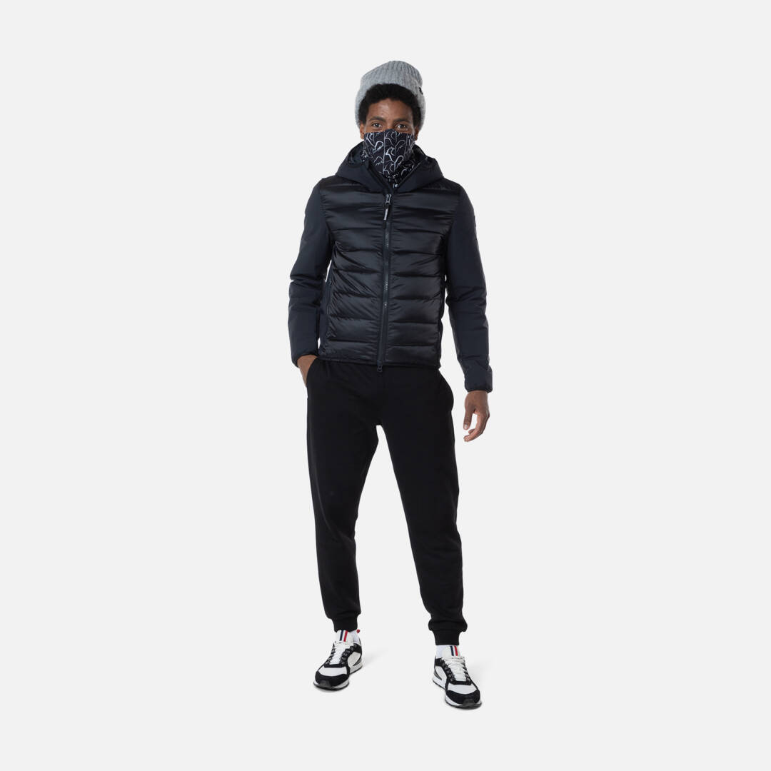 Mann in Rossignol Hoodie Mann mit Mütze und Muster vor Mund, trägt Rossignol Stretch Quilt Hoodie Jacke in Schwarz, Hose, Turnschuhe.