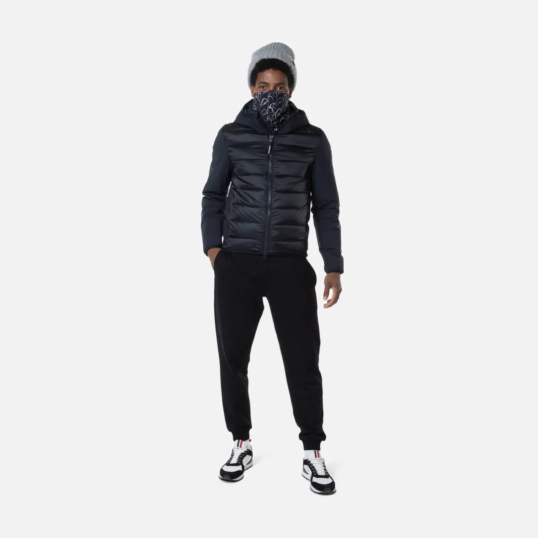 Uomo con berretto e motivo davanti alla bocca, indossa una giacca con cappuccio Rossignol in piumino elasticizzato nero, pantaloni e scarpe da ginnastica.