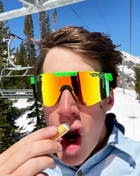 Uomo con occhiali da sole polarizzati verde neon e balsamo per le labbra davanti a un paesaggio montano innevato.