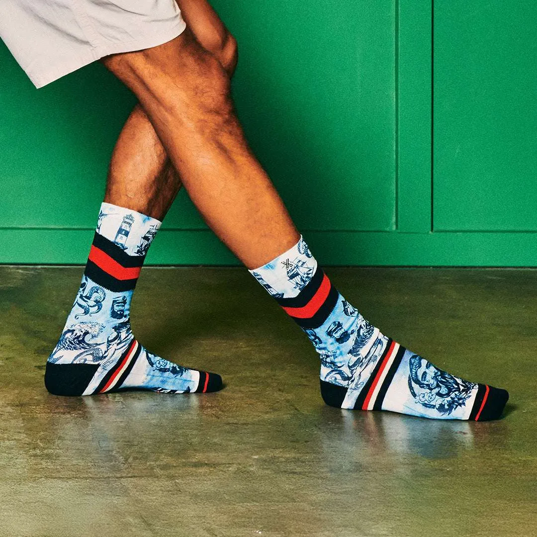 Mann trägt Xpooos Sailor Socken mit maritimen Motiven und roten Streifen vor einer grünen Wand.