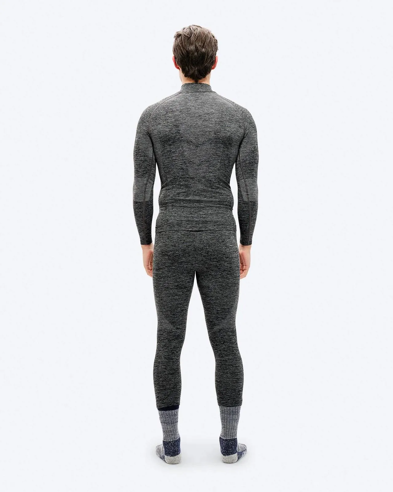 Ein Mann von hinten, gekleidet in ein graues, eng anliegendes, langärmeliges Thermoshirt und eine Hose, trägt dicke Socken. Der Hintergrund ist weiß.