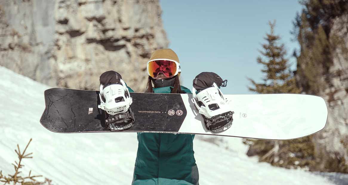 Une personne portant un bonnet et des lunettes de ski tient un snowboard avec des fixations dans un décor de montagne enneigé.