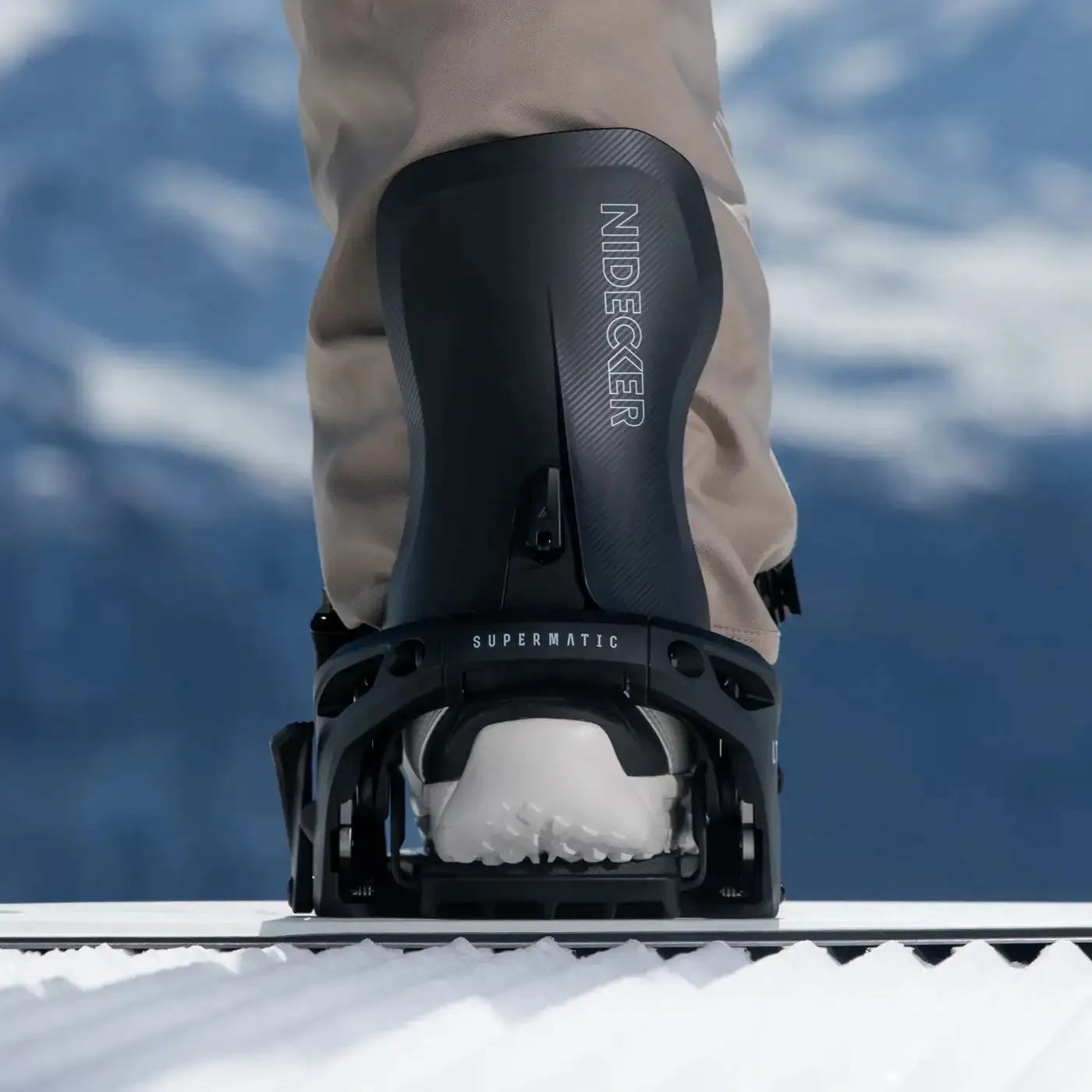 Nahaufnahme einer Snowboardbindung "Nidecker Supermatic" mit Stiefel auf einem Snowboard im Schnee vor verschwommenem Hintergrund.