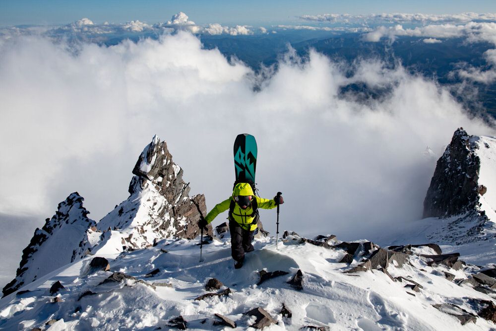 Bergsteiger auf Gipfel Bergsteiger mit Splitboard Jones Womens Solution auf schneebedecktem Gipfel, umgeben von Wolken.