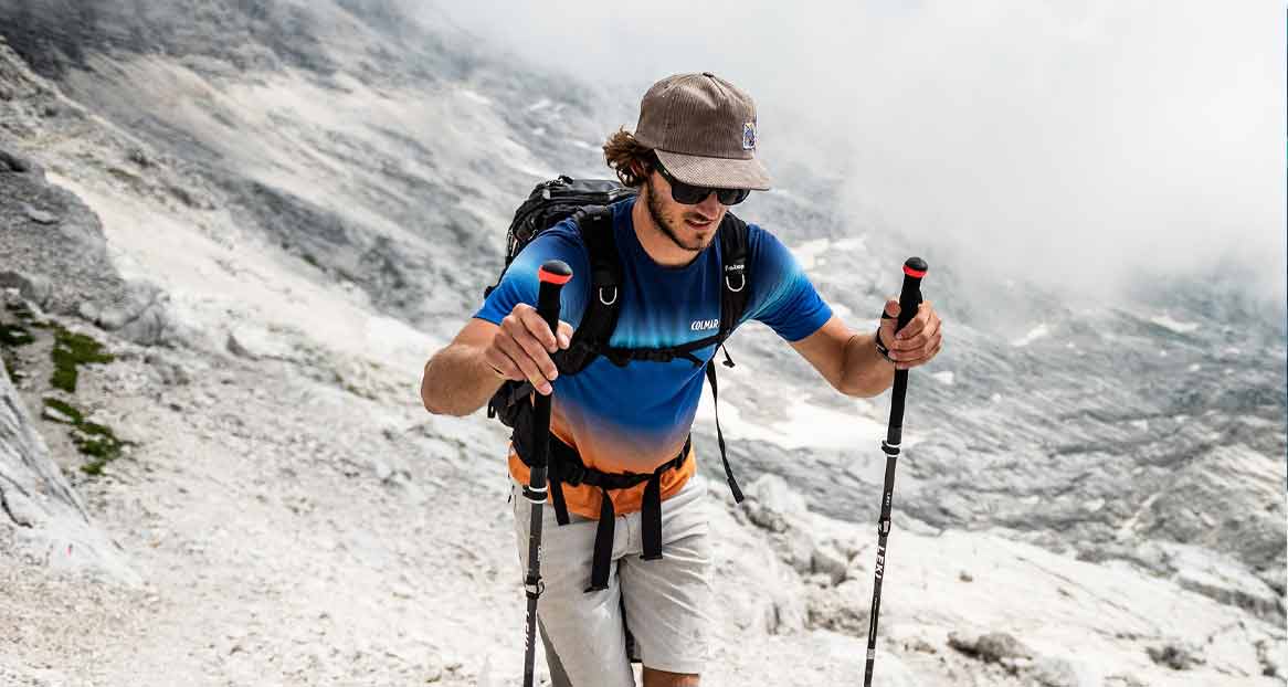 Ein Mann mit Rucksack und Wanderstöcken wandert einen steinigen Bergpfad hinauf. Er trägt eine Kappe und Sonnenbrille.