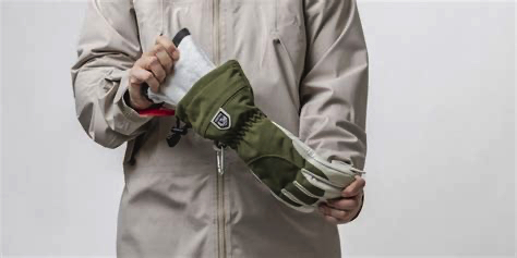 Person, die in einen Armeegrün-Handschuh einen Innenhandschuh steckt. Sie trägt eine beige Jacke.