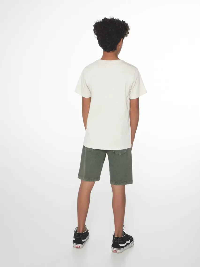 Un enfant aux cheveux bouclés vu de dos, habillé d’un t-shirt couleur crème et d’un short vert olive. Il porte des baskets à motifs noir et blanc. L’enfant se tient devant un fond clair.