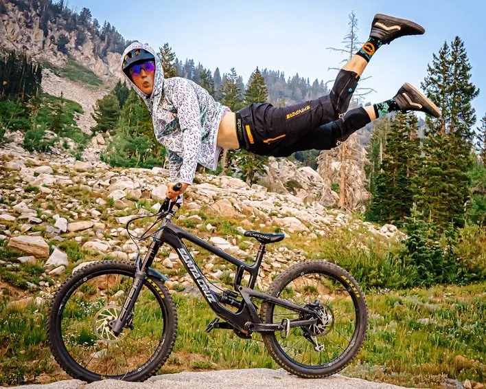 Uomo in equilibrio sulla mountain bike davanti alle montagne. Indossa una giacca con cappuccio, pantaloncini e calzini con un'atmosfera da "afterparty".