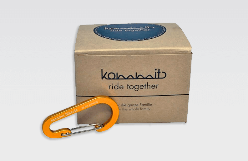 Oranger Karabiner Ride Together Ein oranger kommit Karabiner liegt vor einer braunen Schachtel mit dem Aufdruck "ride together".
