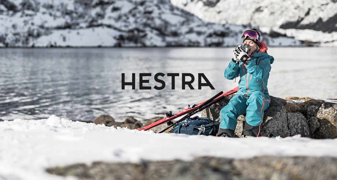 Winter landscape: Woman with ski goggles and ski suit drinking a hot drink by the lake. Skis and rucksack lying in the snow. Hestra logo in the foreground.