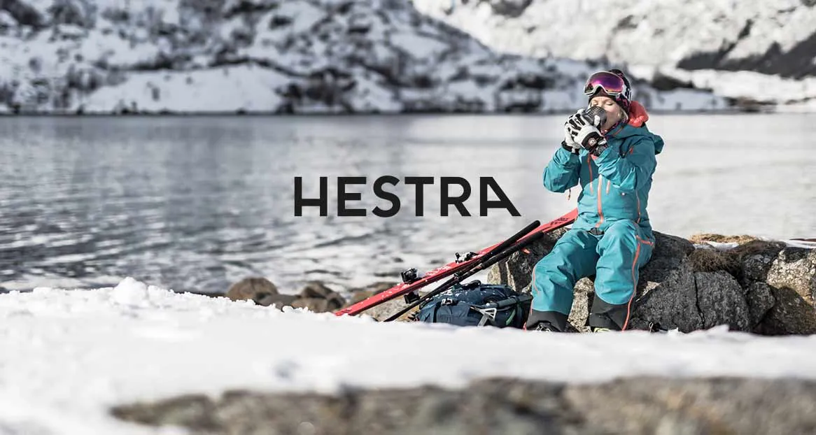 Winterlandschaft: Frau mit Skibrille und Skianzug trinkt Heißgetränk am See. Ski und Rucksack liegen im Schnee. HESTRA-Logo im Vordergrund.